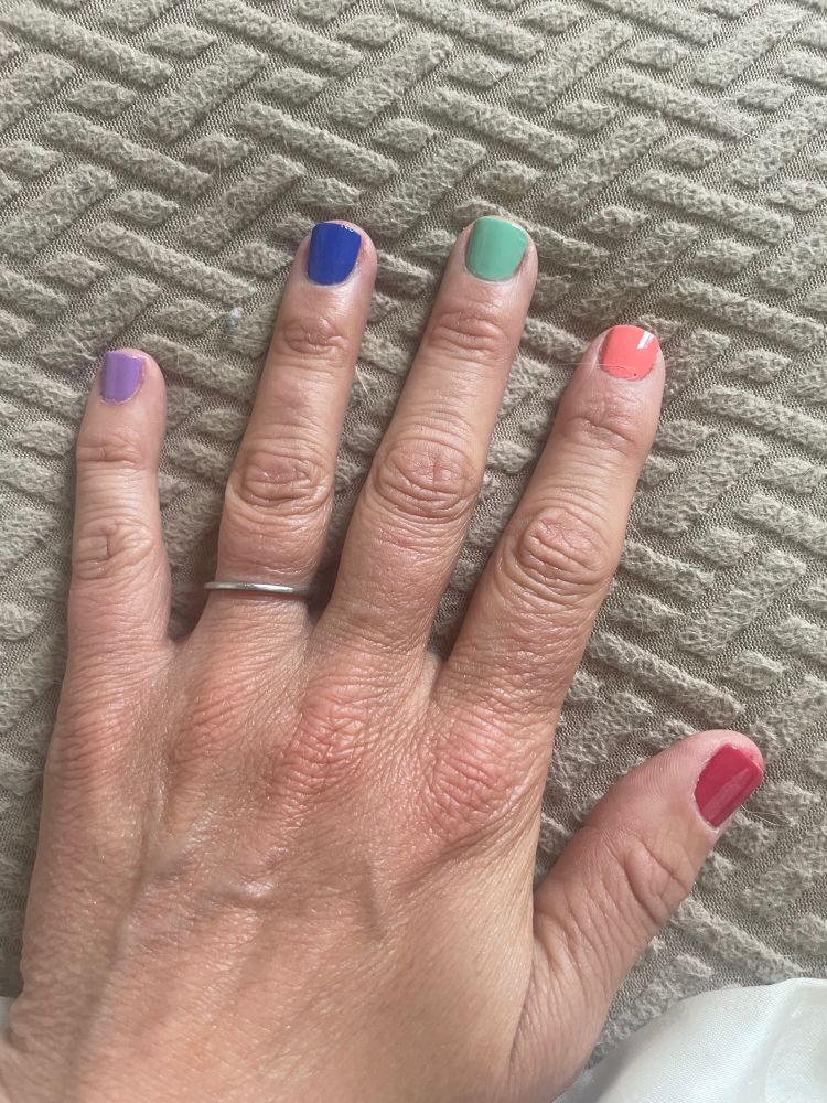 My left hand with pride manicure. Fingernails each painted a different color to make a rainbow. Thumb is dark pink, pointer is peach, middle is green, ring is blue and pinky is purple. I’m wearing a thin platinum wedding ring on my ring finger.