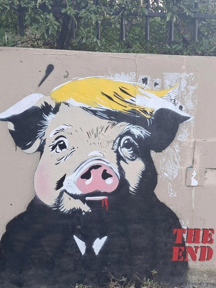 graffiti art on a wall, artist unknown. 
The art depicts a pig dressed a Donald trump in a cartoon style. The pig is wearing a Black suit and tie, with yellow toupee hair falling to the right side. small red drips are seen from the right side of the mouth and at the bottom right of the wall is text in red that reads "THE END"