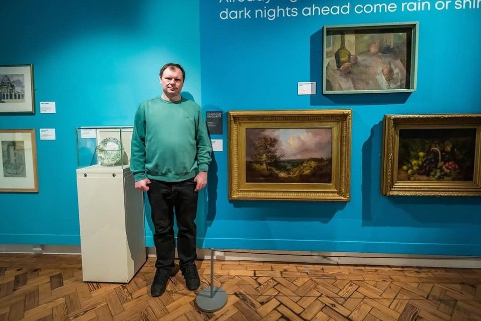 A person in a green sweater stands in an art gallery. They're next to a blue wall displaying various framed paintings and art pieces. The floor has a wooden parquet pattern, and a sign above reads, "dark nights ahead come rain or shine.