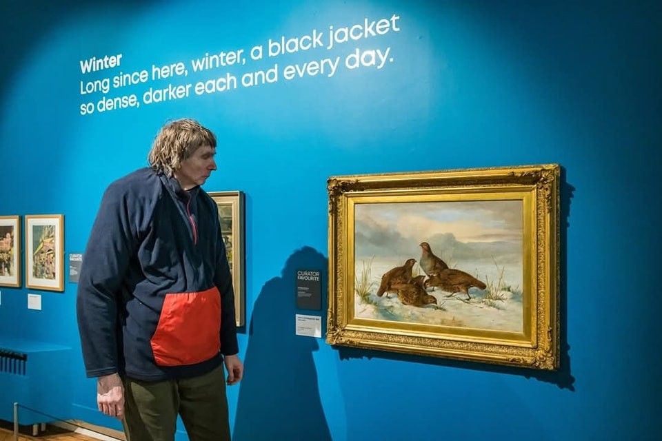 A person wearing a black and red jacket observes a painting in a gallery. The blue wall displays a poem titled "Winter." The painting in a gold frame depicts three birds in a snowy landscape.