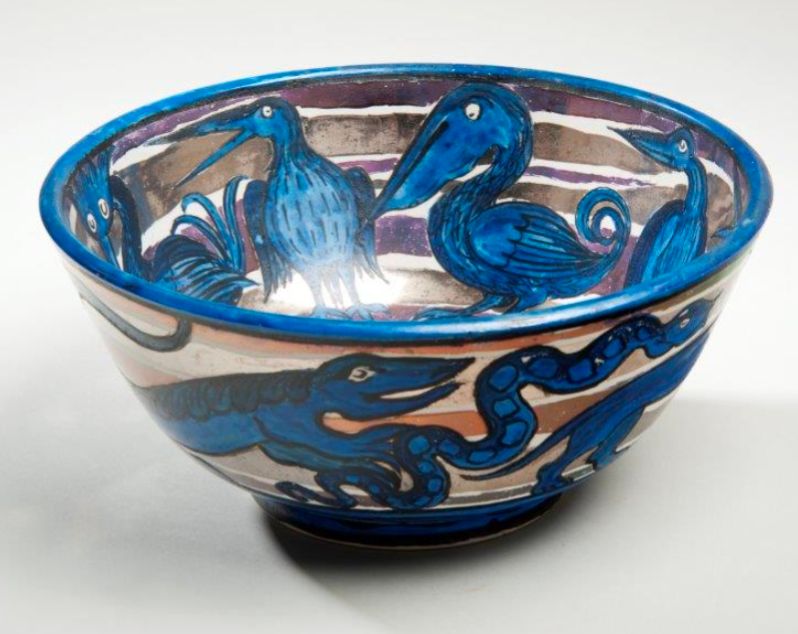 A ceramic bowl finished with bands of lustre glazes and stylised birds in blue. In the centre of the bowl is a peacock and around the outside are various animals including a snake, lizard, wolf and crocodile.