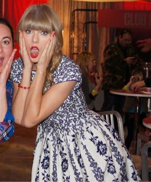 Taylor Swift wearing a white dress with blue floral patterns, poses with her hands on her cheeks in surprise. She's indoors at an event with people and tables in the background.