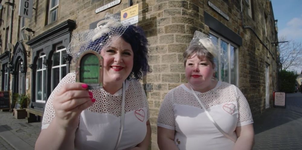 The Brides of Eldon Street, Gemma and Beck in wedding dresses, holding a small green door to the camera.