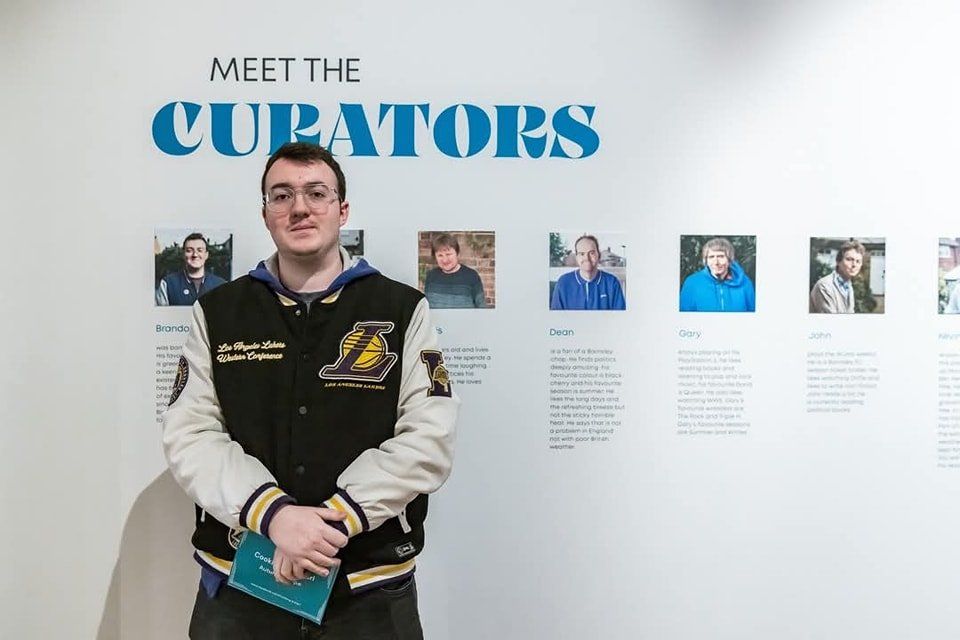 A person wearing a Los Angeles Lakers jacket stands in front of a wall displaying "Meet the Curators" with photos and descriptions of various individuals. They hold a booklet and appear to be in an exhibition or gallery setting.