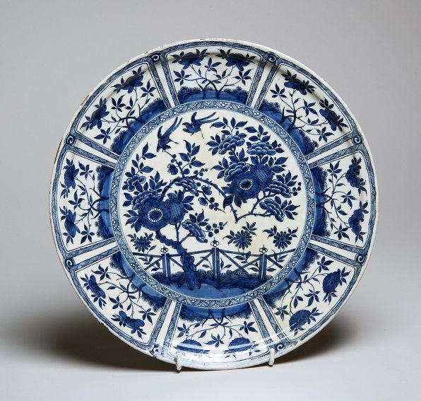 A decorative porcelain plate featuring intricate blue floral and bird designs on a white background. The border has panels with leaf motifs. The plate is displayed upright on a small stand.