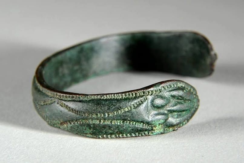 A close-up of an ancient, greenish metal bracelet with an open end, featuring engraved decorative patterns along its surface and a slightly weathered texture.
