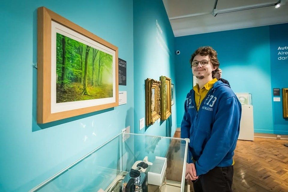A person in a blue hoodie and glasses stands smiling in an art gallery. They are next to a framed painting of a vibrant green forest on a teal wall. The gallery has various artworks and a display case on a wooden floor.