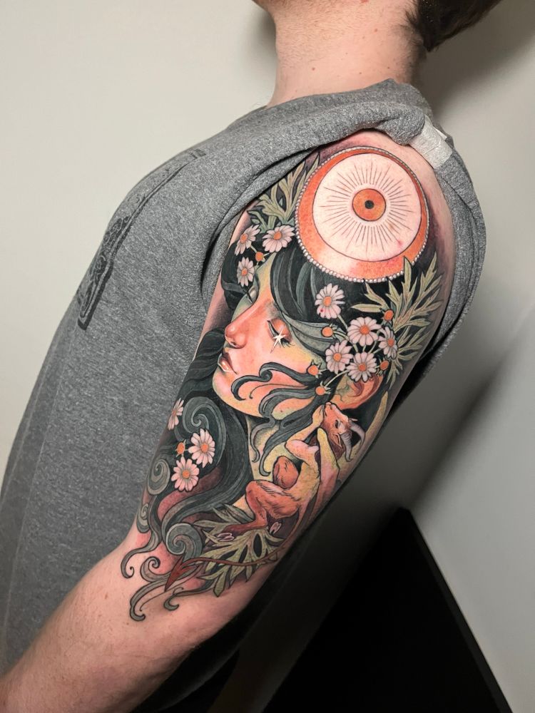 Tattoo on a mans upper arm of a lady with dark hair. She is surrounded by swirls of far hair and camomile flowers. A tiny goat man imp sits on her shoulder and whispers to her