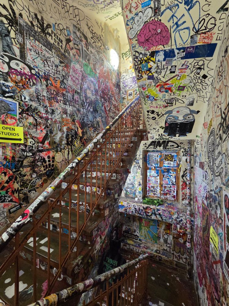 Ein Treppenaufgang mit Graffiti an den Wänden, an der Decke, auf dem Fenster und auf der Treppe. Links ein Hinweisschild: open studios.