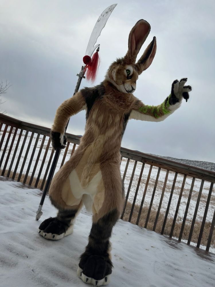 A brown jackrabbit fursuiter wielding a guandao strikes a pose, as if he were ready to challenge an opponent offscreen. The sky is grey and the landscape is a snowy winter scene.