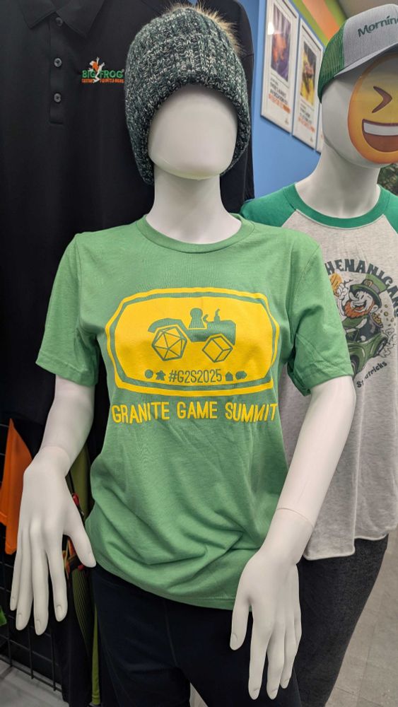 A green shirt with a yellow picture of a tractor with piece components and the words Granite Game Summit below that on a mannequin doing what I can only assume is lower Thriller arms. 
