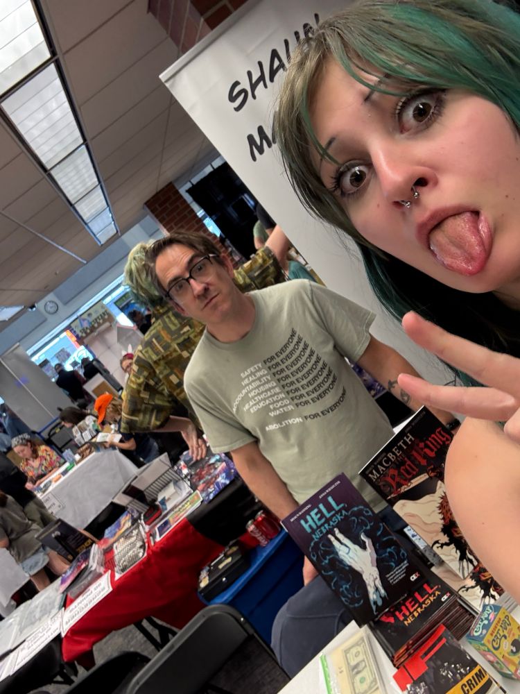 Ray, a teen girl with green hair, sticks out her tongue in the foreground while Shaun stands at the table