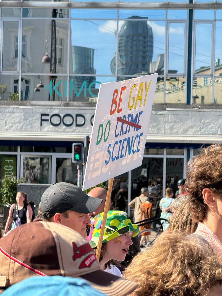 A parade member with a green bucket hat holding a sign that says Be Gay Do Crime, with the Crime slashed out and Science written underneath