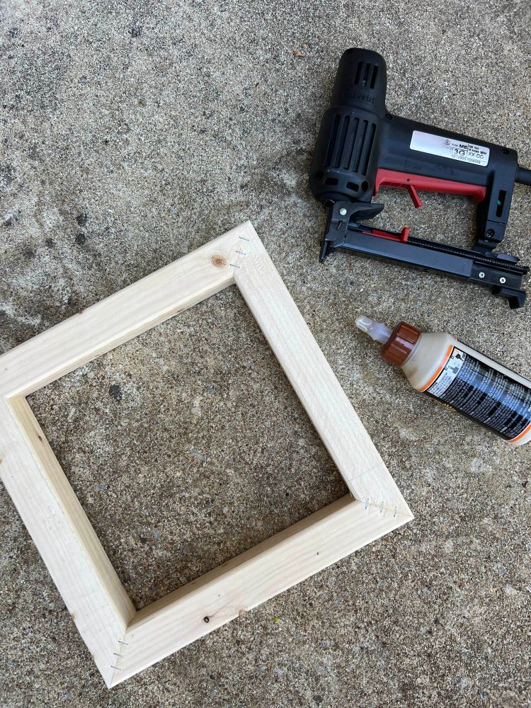 A small square wooden frame sits on concrete next to a bottle of wood glue and a staple gun.

