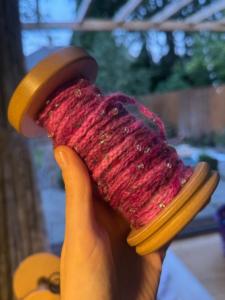 My hand is holding a wooden spinning wheel bobbin 3/4s full of bright pink chainette yarn with silver glass beads spaced evenly on it. 
