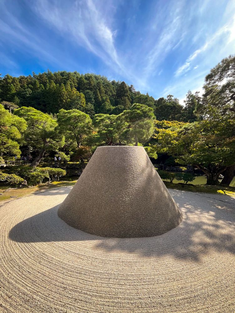 Un jardín zen (gravilla rastrillada) formando círculos alrededor de un cono de piedra sin pinta que recuerda vagamente el monte Fuji. Detrás, los árboles del jardín.