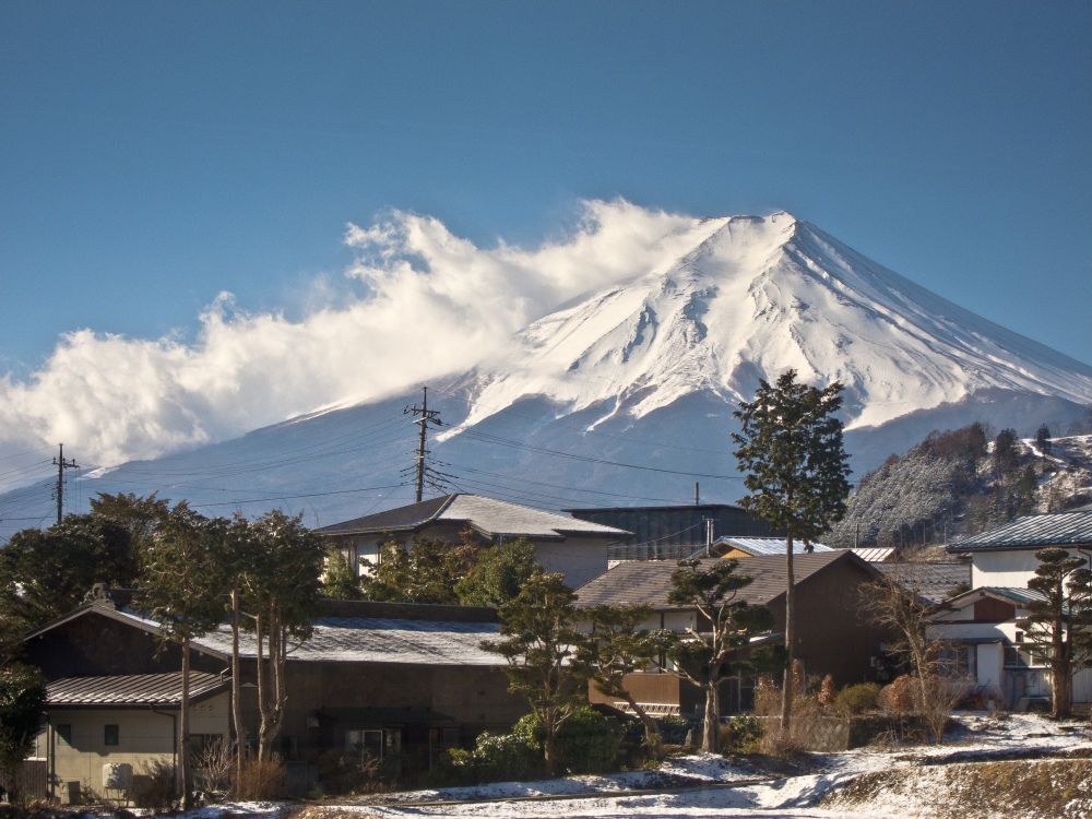 Vista del Fuji con el cono superior nevado y una "cresta" de nubes a su izquierda. En primer plano, casas rodeadas de unos pocos árboles y nieve en el suelo y los tejados.