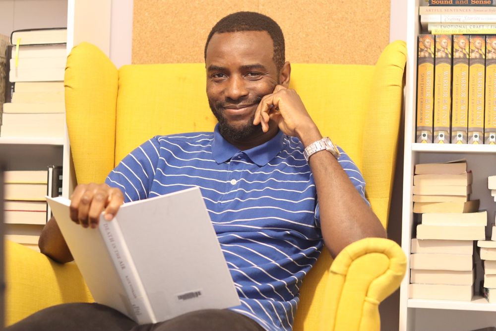 Author Suyi Davies Okungbowa, in a blue shirt striped with white, sits on a yellow armchair, with bookshelves in the background, reading a white hardcover book and smiling at the camera.
