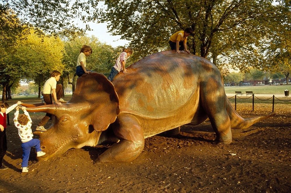 A fiberglass statue of a triceratops, Uncle Beazley, at the National Mall in Washington, D.C. The statue has a copper and sort of verdigris patina. Young children are climbing the statue. Its a Fall day, there are trees in the background.