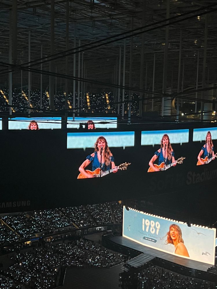 Taylor Swift on projector screens at Sofi Stadium holding a guitar and smiling wearing a blue off the shoulder dress with 1989 Taylor’s Version on the main screen. 