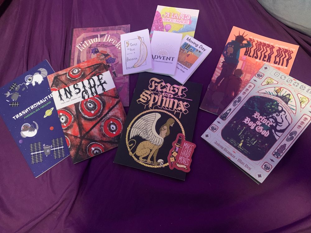 A selection of ttrpg zines on a purple sheet. Titles include ‘Before the Bog God, A Land Once Magic, Inside Out, Feast for a Sphinx, Blister City, Ritual Deck, 5 Games to Play With a Banana, Advent, and Moving Day.’