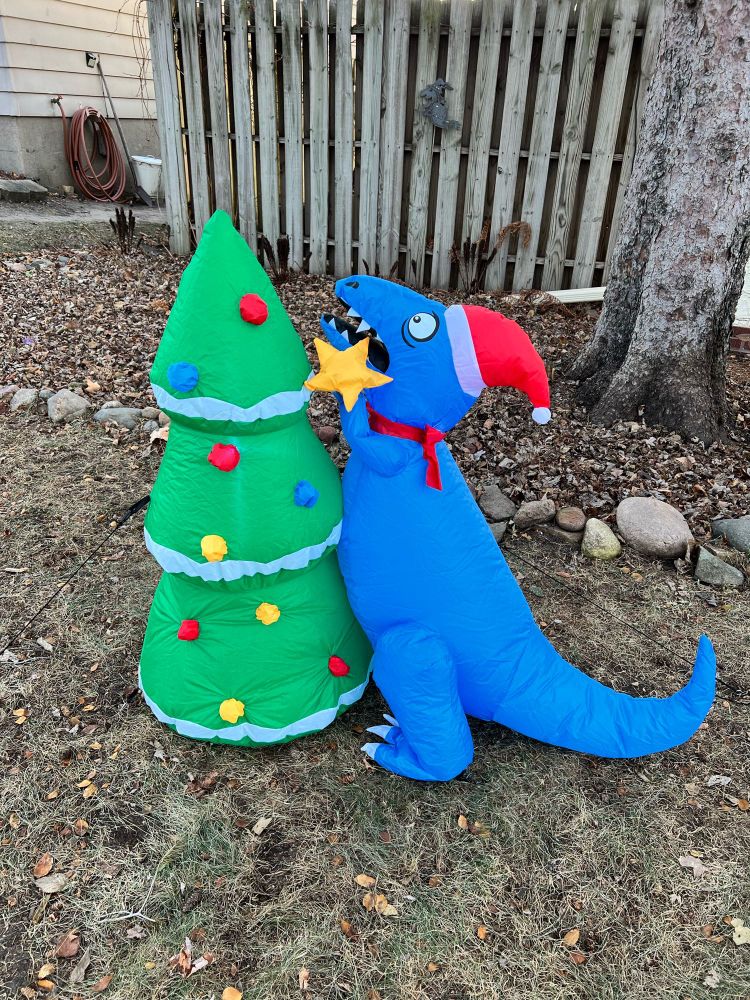 Outdoor blow up decoration of a T-Rex trying to put a star on top of a Christmas tree but cannot reach
