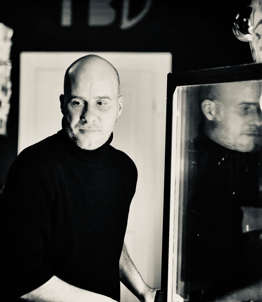 Schwarzweiß Brustporträt von mir hinter der Bar. Ich trage einen schwarzen Rolli und kein Cap