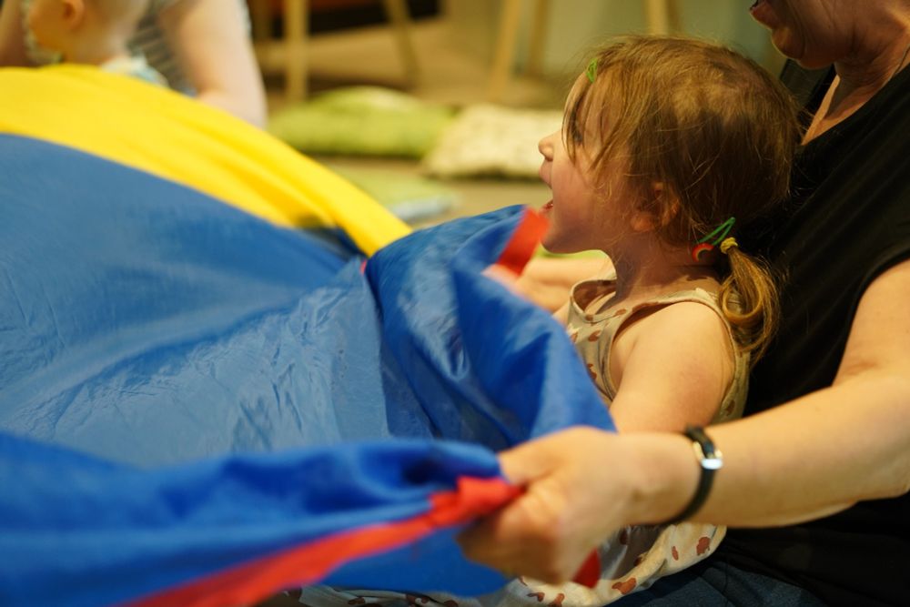 A small girl is smiling and laughing, while a rainbow patterned sheet is waved around her. 