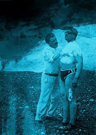 Two men stand on a pebble beach. The man on the right is wearing speedos and goggles, being covered in goose fat by the man on the left.