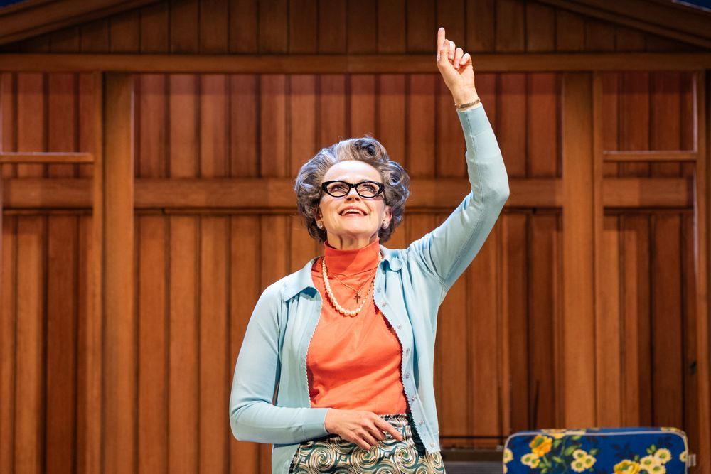 A garden shed against a blue backdrop on a stage. Standing in front of it a woman wearing a blue cardigan, pink rollneck and patterned skirt. She is pointing one finger up and looking upwards with a smile on her face.