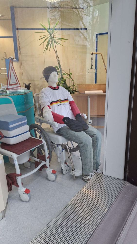 Schaufensterpuppe in Schaufenster gekleidet in Deutschlandtrikot und wilder Mütze, heißer drip 🔥.  Sitzt in einem ergonomischen Rollstuhl für Menschen mit Behinderung. 