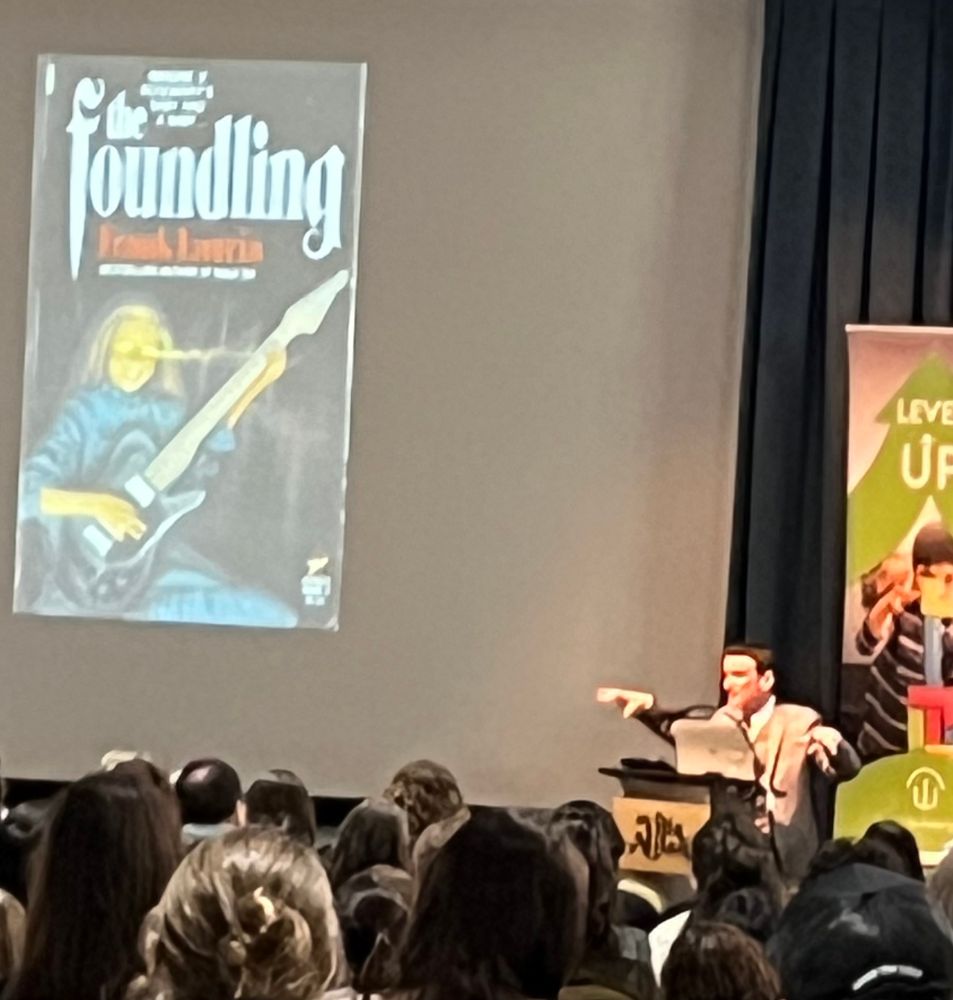 Author Grady Hendrix at podium with arms dramatically in the air while a vintage book cover for the Foundling is projected on the screen next to him.