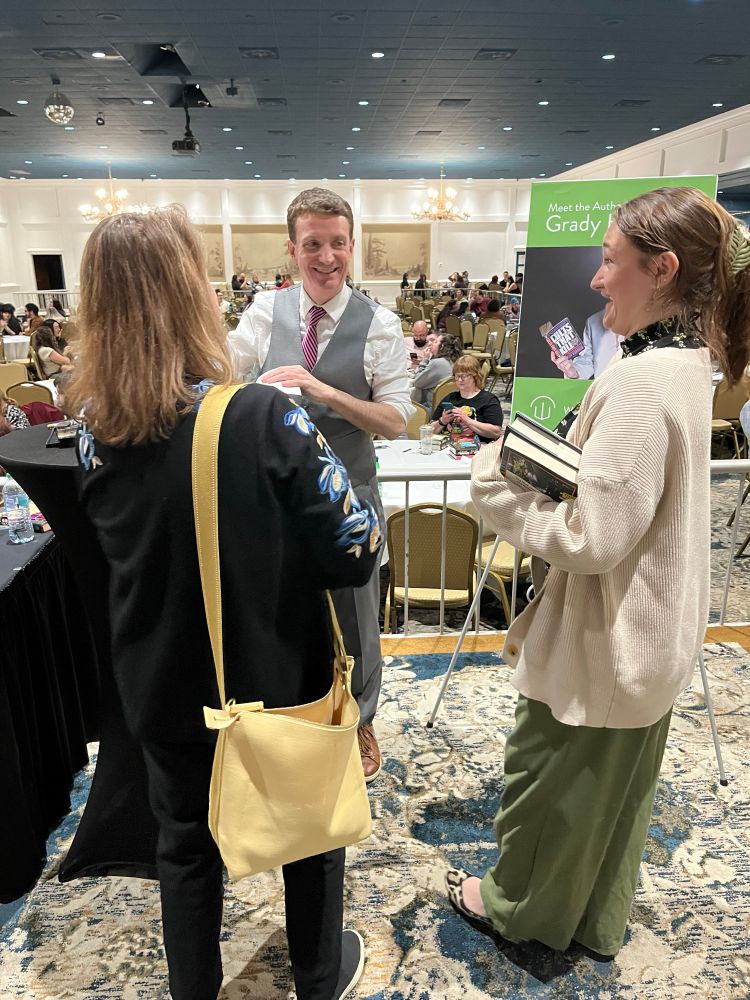 Author Grady Hendrix smiles as he signs books for mother with back to camera and young adult daughter who is smiling as she chats with him.
