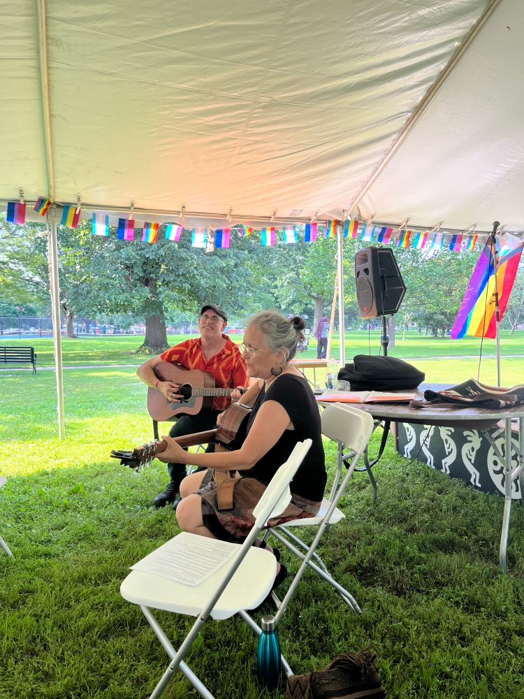 Man in red shirt with guitar smiling at woman in black shirt with guitar sitting next to him and singing. Pride flags in the background.