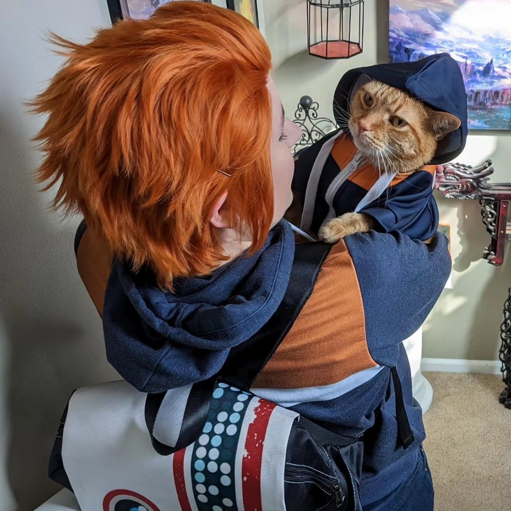 Photo of me wearing my (unstyled, lol) Cal wig, plus hoodie and BD-1 backpack, holding Brady, a big orange tabby cat wearing a tiny kitty version of the Cal hoodie. The hood is up in this photo and one ear is poking out