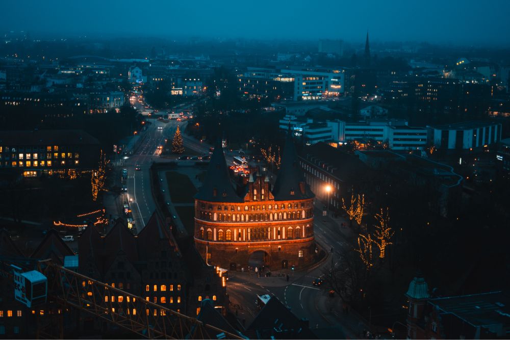 Old city gate illuminated at night, around swirls a road, blue night sky behind it. Viewed at an elevated angle from a church tower