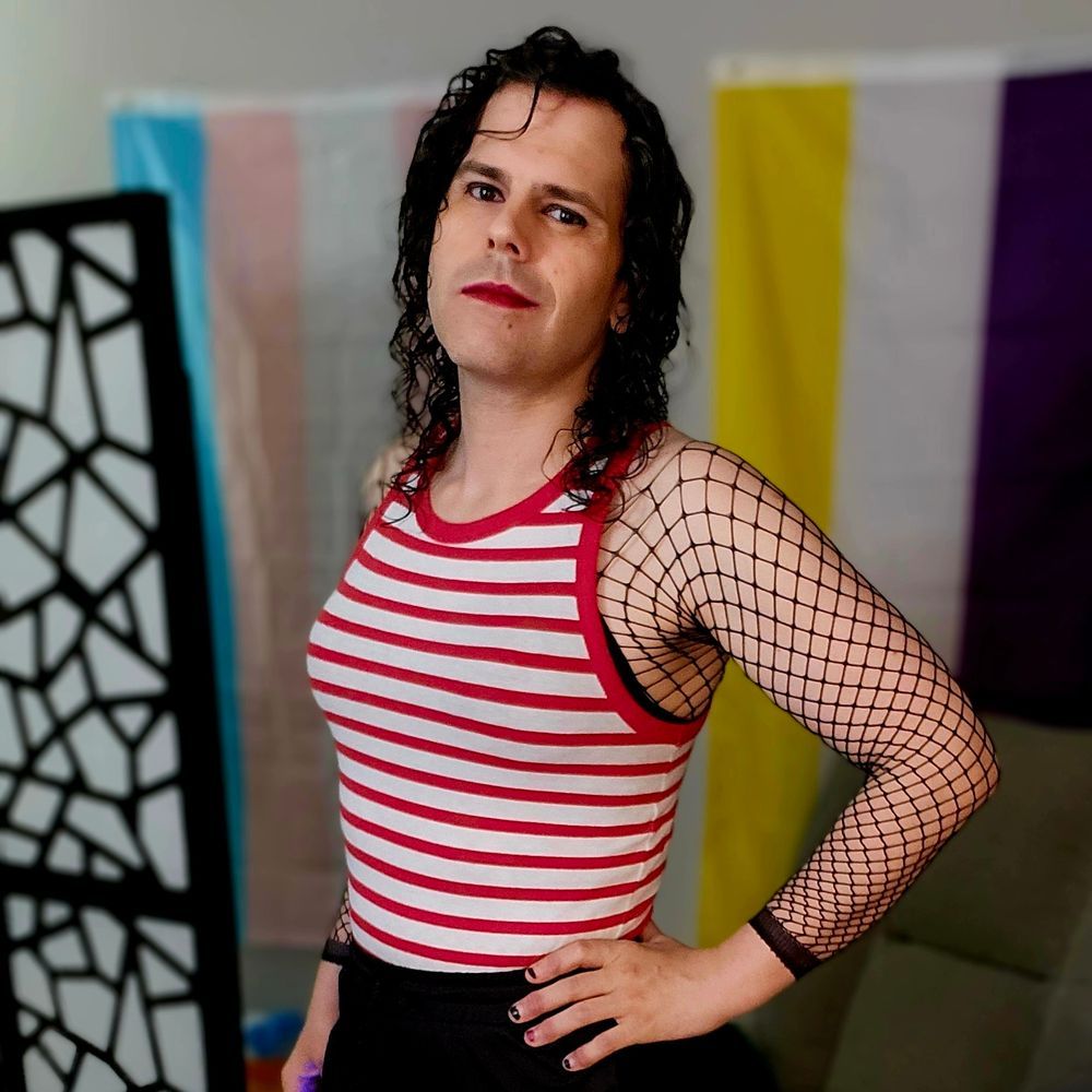 Zoe in red and white horizontal striped shirt with black fishnets for sleeves. Her hair is a deep black and her eyes a dark brown. She has deep red lipstick and curly shoulder length hair. Her hand is on her hip revealing 4 black fingernails and one red on her ring finger. The background is out of focus but a trans and nonbinary flag can both be made out.