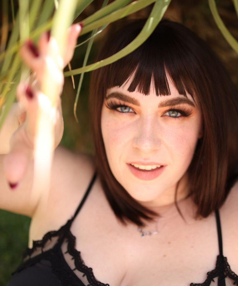 A close up of Jane. She has brown hair, long lashes, blue eyes, and pink lips. She is wearing a silver necklace, and a black top, with her right hand visible showing her long red fingernails.