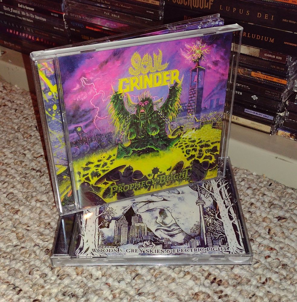 the CD case for The Prophecy of Blight by Soul Grinder sitting on top of the CD case for Woods 5: Grey Skies & Electric Light by Woods of Ypres