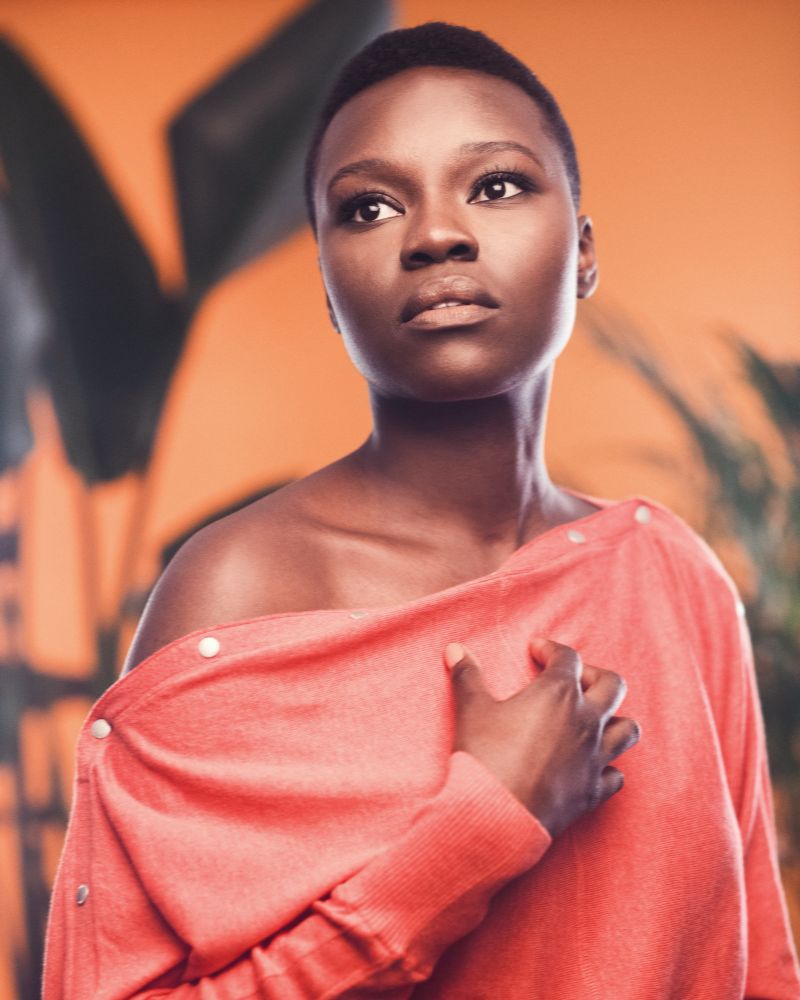 A photo of Mina from The Resident in an orange off the shoulder dress with her hand on her chest looking away from the camera 
