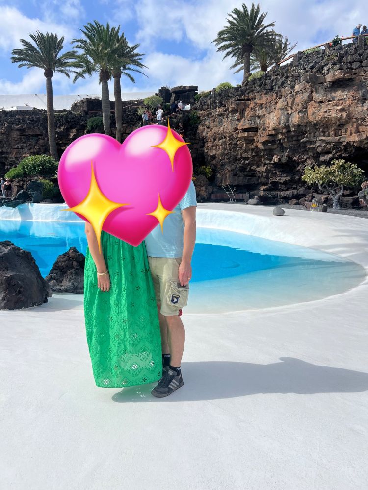 Bild von uns vor Manriques Poollandschaft. Unsere Oberkörper sind von einem Herzen verdeckt - der Gatte ist nicht scharf auf Social Media - Präsenz. Ich trage ein grünes Kleid mit Lochstickerei, der Gatte ein türkisfarbenes Shirt, beigefarbene Bermudas, dunkle Socken und Sneakers. Es wirkt, als schwebe ich, da man meine Füße nicht sieht, obwohl das Kleid nicht bodenlang ist.