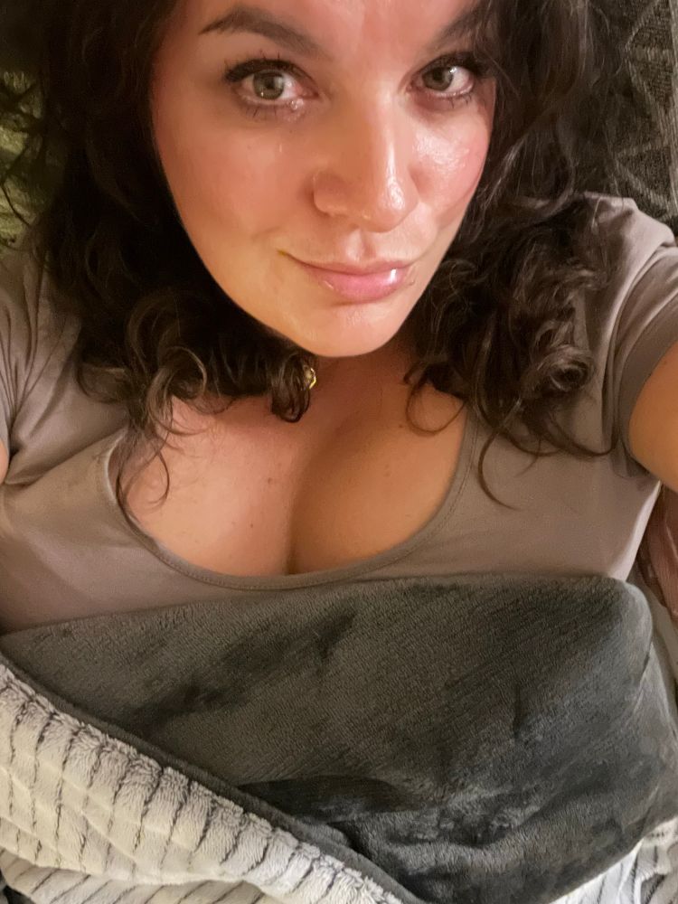 Auf dem Bild liegt eine Frau auf einer weichen Unterlage und blickt direkt in die Kamera. Ihr Gesicht ist nah aufgenommen, sodass ihre Augen und ihr Ausdruck gut zu erkennen sind. Sie hat dunkle, leicht lockige Haare, die über ihre Schultern fallen.

Sie trägt ein kurzärmliges Oberteil in einem hellen Braun- oder Beigeton. Über ihrem Körper liegt eine zusammengerollte Decke in Grau und Weiß. Der Hintergrund wirkt gemütlich und besteht aus weichen Stoffen, was den Eindruck eines entspannten Moments vermittelt.

Die Beleuchtung ist warm und lässt ihre Haut sanft wirken. Der Gesamteindruck des Fotos ist persönlich und ruhig.