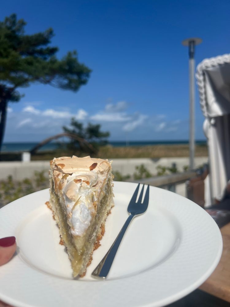 Ein Stück Stachelbeerbaisertorte auf einem Teller mit Gabel. Im Hibtergrund ein Strandkorb und noch dahinter die blaue Ostsee und ein Windflüchter.