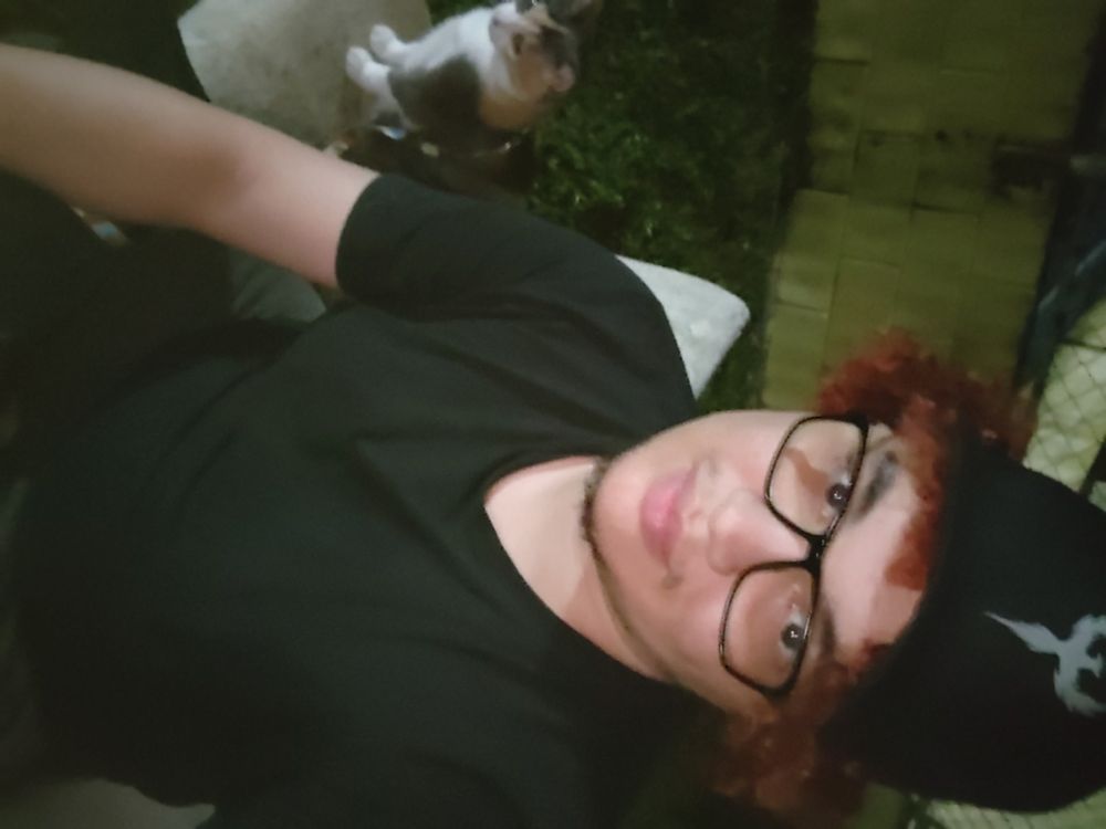 É noite. Sentado num banco de pedra estou vestindo camiseta, calça e boné pretos. encaro docil a câmera. Mechas vermelhar saem do boné. Ao fundo grama e um muro amarelado. Também no banco, com olhar vago, um gato tricolor está com postura sentada.