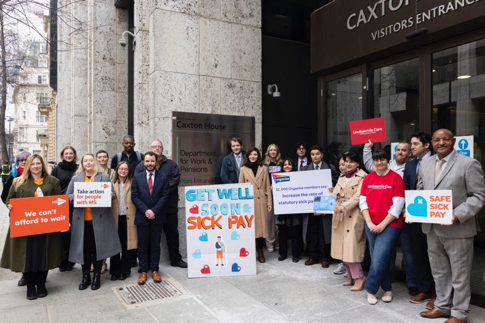 Campaigners stand outside entrance to Department for Work & Pensions with the Giant get well soon card, and signs from MS Society, Leukeamia Care and Organise. Credit: Sylvie Pope Photography, Safe Sick Pay campaign