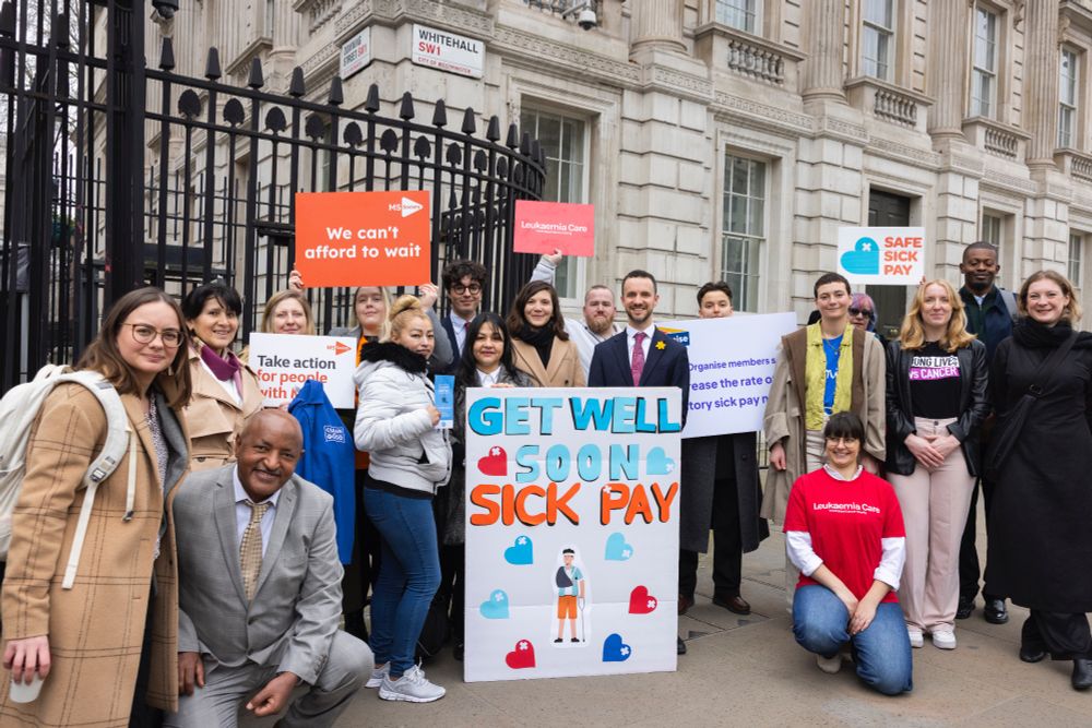 Campaigners stand outside gates to Number 10 Downing Street with the Giant get well soon card, and signs from MS Society, Leukeamia Care and Organise. Credit: Sylvie Pope Photography, Safe Sick Pay campaign