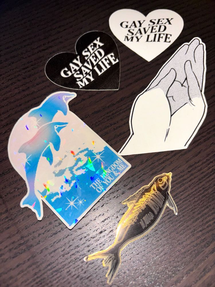 Selection of 5 stickers. There's a black heart & white heart, both saying "Gay Sex Saved My Life", a hand in the shape of a fist, a holographic window with two dolphins leaping out of pixellated water with the caption "the kingdom of you and me", and a shiny gold fish skeleton.