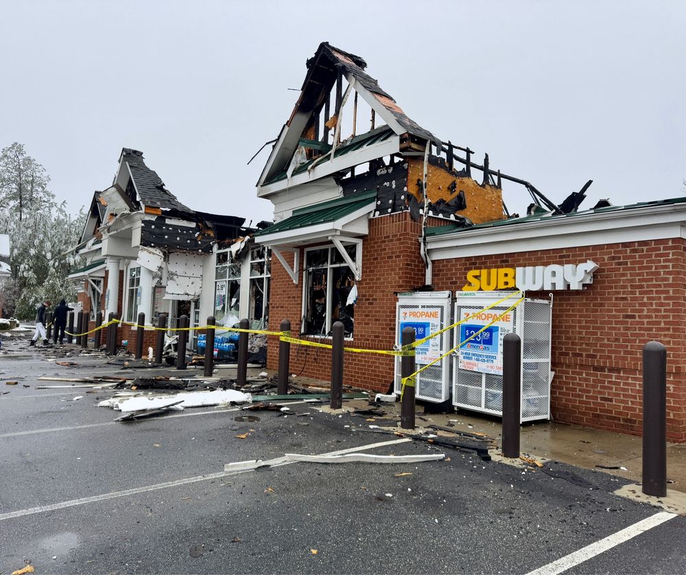 The front of a burned gas station building with a subway sign and propane tanks in the forefront. Yellow caution tape wrapped around poles in front of it. 