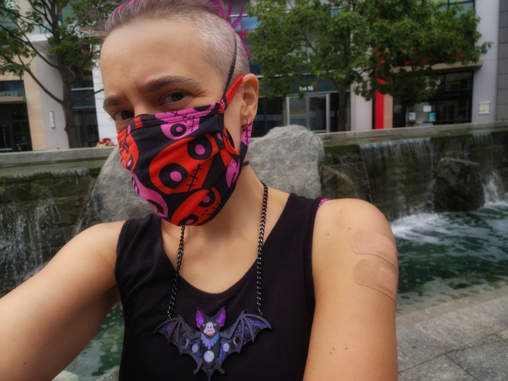 Selfie outside the CVS where I just got vaccinated, in front of some waters feature with rocks and falling water. I'm showing off two bandaids on my left shoulder. I'm wearinga black sleeveless tunic, a black face mask with pitch and red cartoon skulls on it, and a large acrylic bat pendant in a sparkly black and purple with moon phases on the body and outstretched wings.