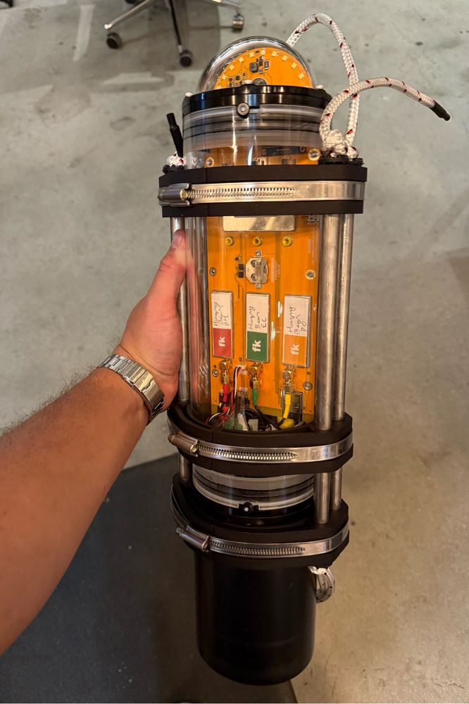 A hand holds out a fully assembled FieldKit.org underwater sensor sitting on a table. The unit measure pH, conductivity, temperature, depth, and dissolved oxygen. 